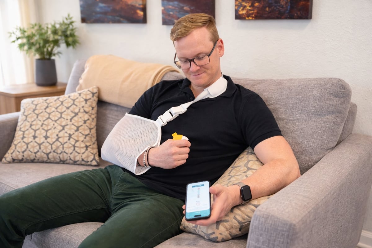 Man sitting on gray couch with arm in cast, holding smartphone and snack, wearing glasses and casual attire in modern living room