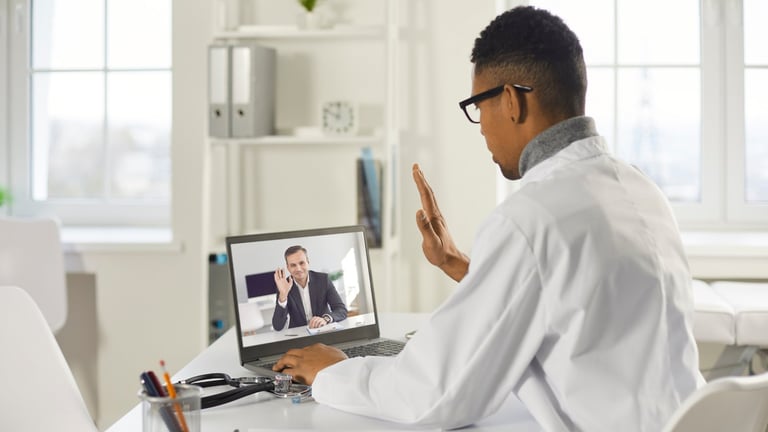 Man on video call waving at laptop screen during a virtual meeting in an office setting