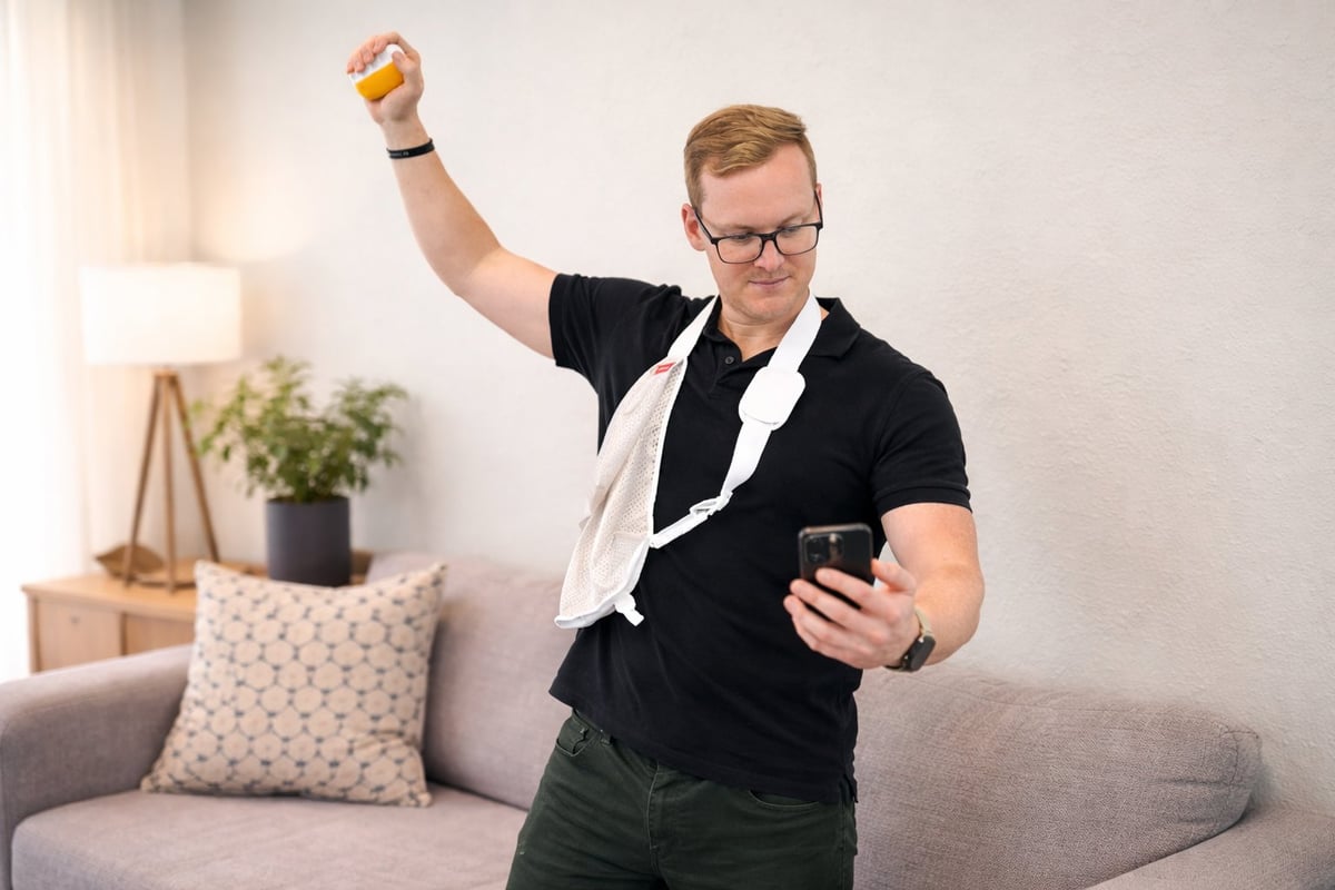 Man in glasses and black shirt holding phone with fist raised in celebration, standing in front of gray couch with white towel and plant